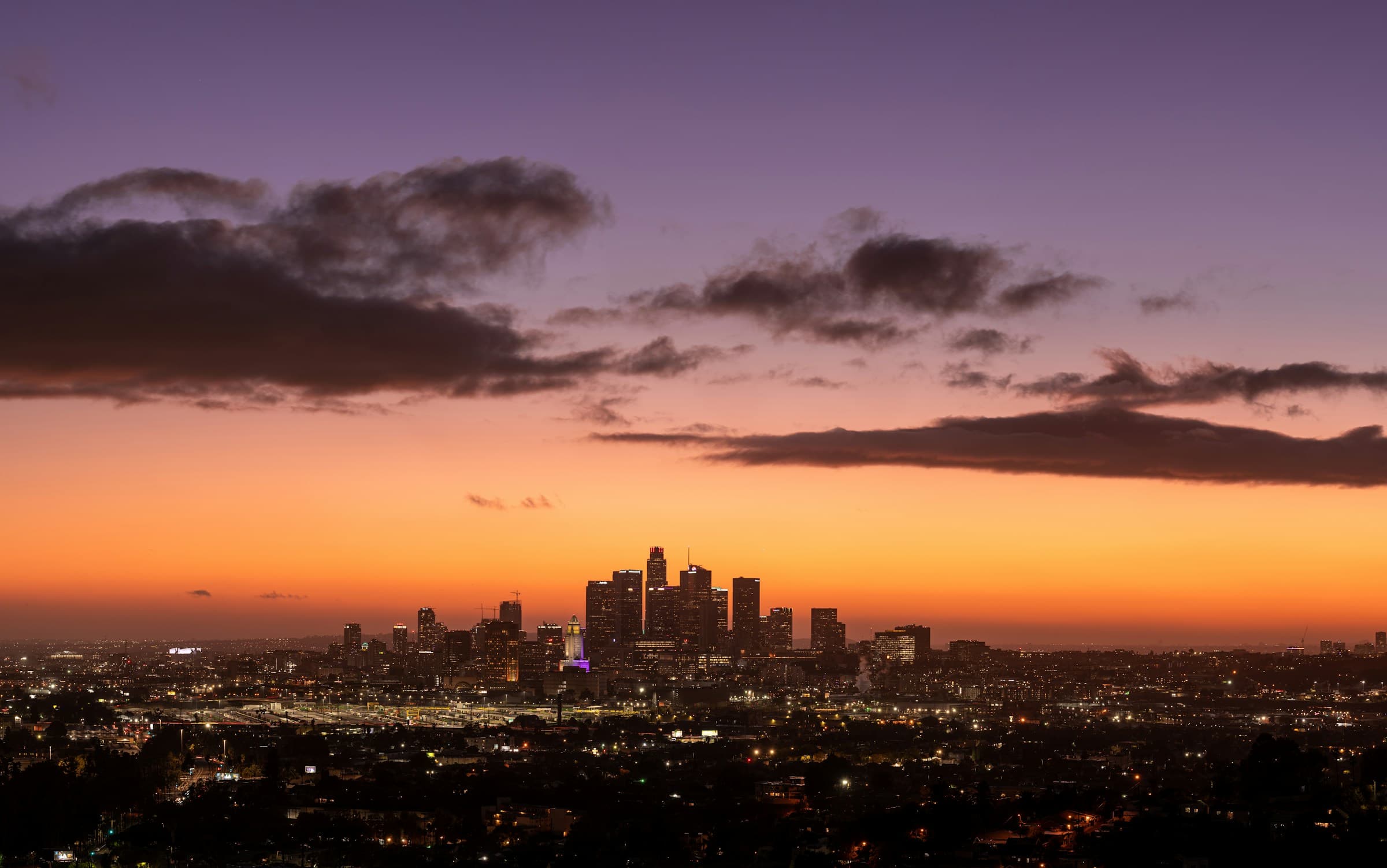 The downtown Los Angeles skyline silhouetted against an orange and purple sunset, dramatic clouds overhead, city lights spreading across the LA basin below.